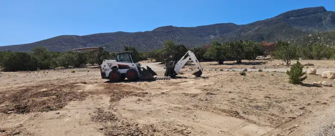 A Bobcat excavator digging a trench in Placitas, New Mexico, preparing the ground for a concrete workshop foundation.
